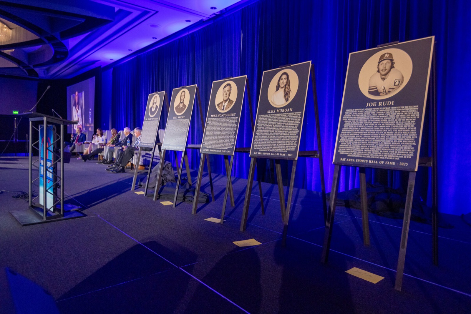 Row of BASHOF 2025 inductee plaques on stage, including Dr. Harry Edwards, Eric Wright, Mike Montgomery, Alex Morgan and Joe Rudi